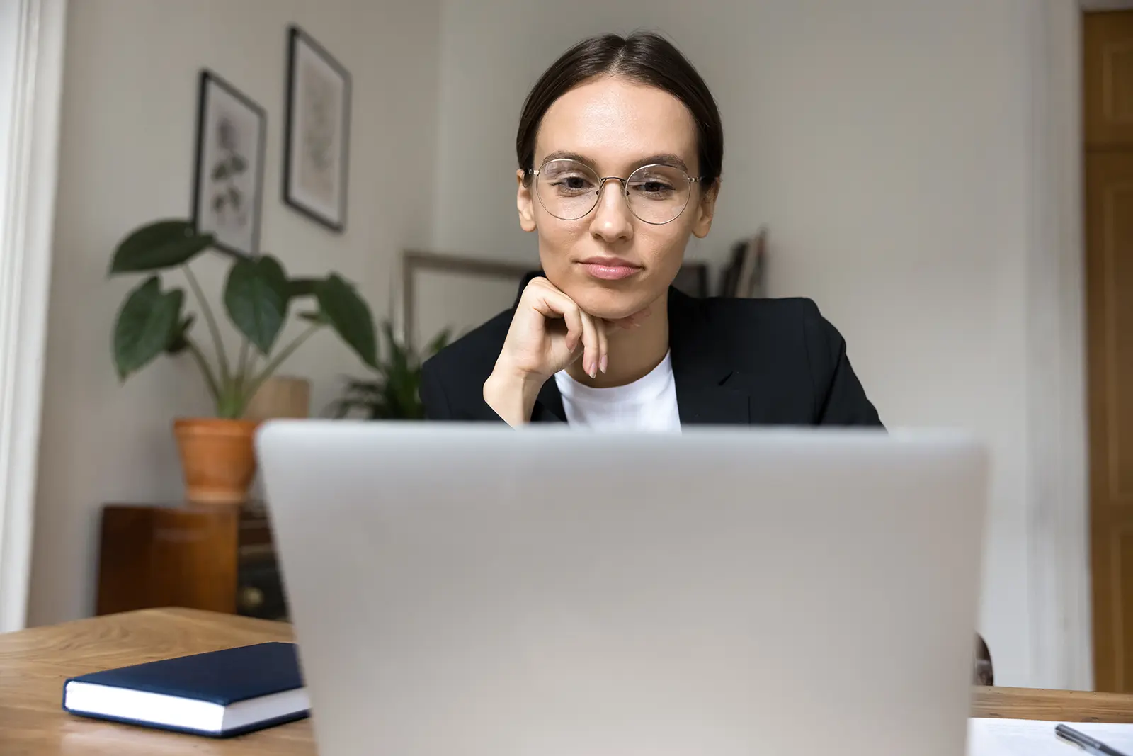 Giovane donna d'affari positiva, concentrata e riflessiva che usa la comunicazione online per lavoro, lavora al computer portatile in ufficio a casa, toccandosi il mento, guardando il computer attraverso occhiali alla moda, pensando al font giusto da usare sul suo sito